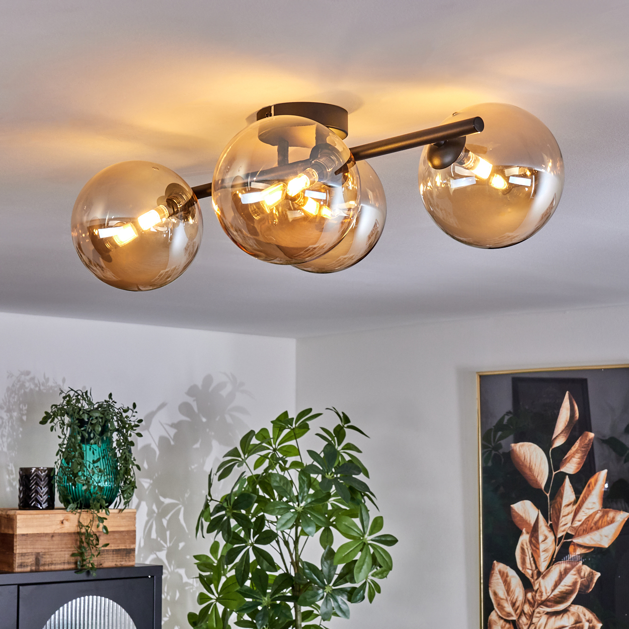 Deckenlampe mit drei goldenen, kugelförmigen Lichtern in einem Raum. Pflanzen und Möbel befinden sich im Hintergrund.