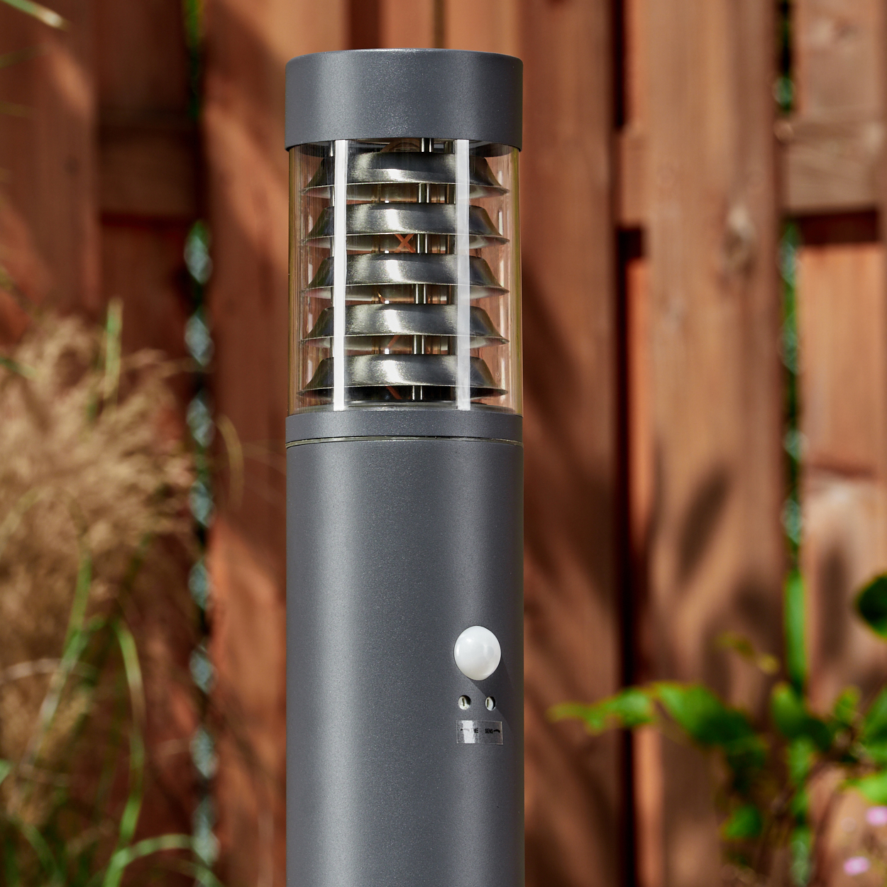 Nahaufnahme einer grauen Außenlampe mit Bewegungssensor, mit verschwommenem Gartenhintergrund.