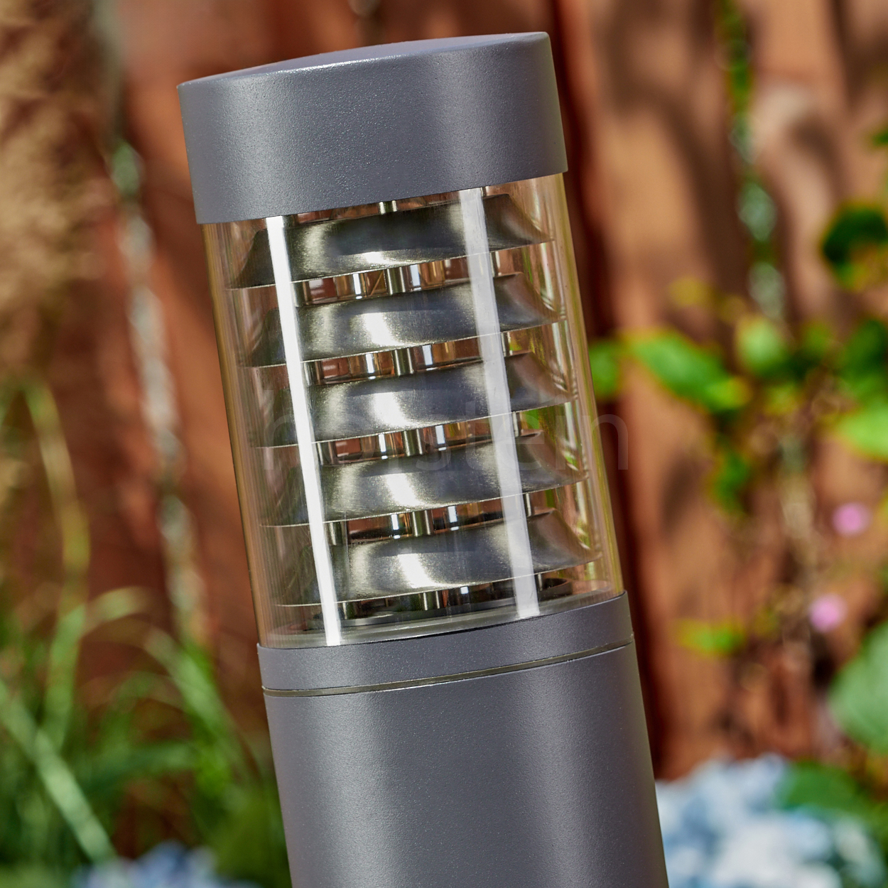 Nahaufnahme des oberen Teils einer grauen Außenlampe, mit unscharfem Gartenhintergrund.