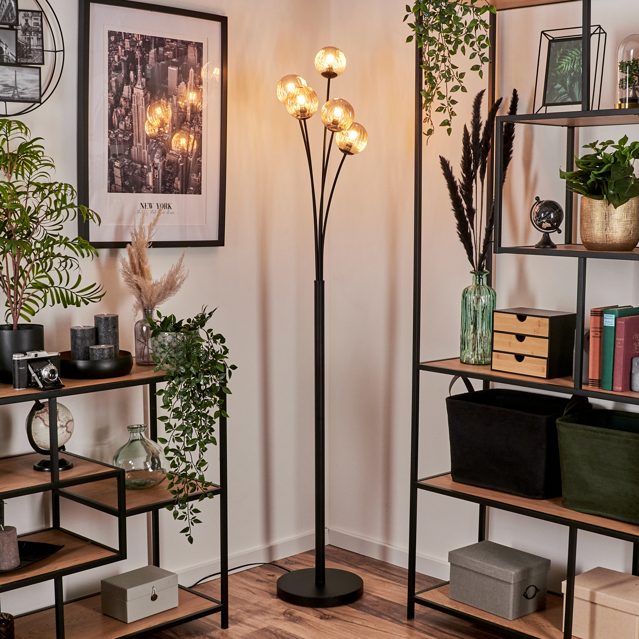 Eine Stehlampe mit bernsteinfarbenen Kugeln steht in einem hellen Raum mit Regalen, Pflanzen und einem gerahmten New York City-Bild.