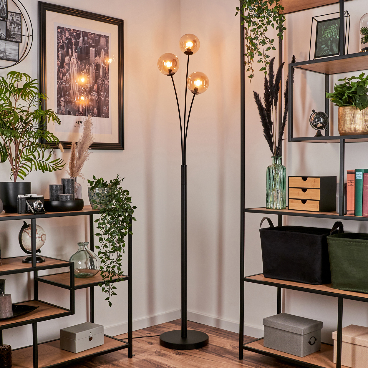 Schwarze Stehlampe mit drei bernsteinfarbenen Kugeln. Raum mit Regalen, Pflanzen und einem gerahmten Bild im Hintergrund.