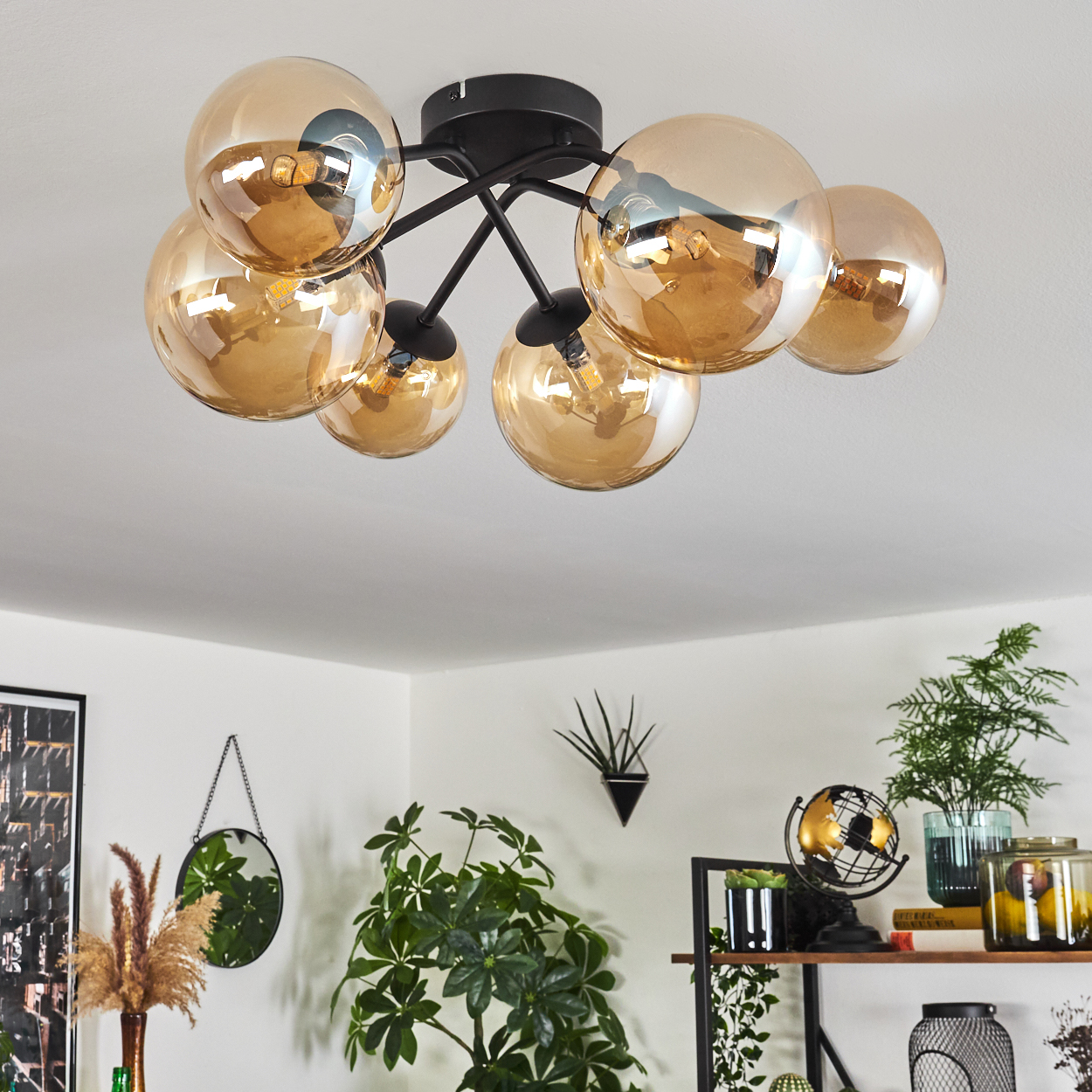 Deckenlampe mit bernsteinfarbenen Glaskugeln, schwarzer Rahmen. Innenraum mit Pflanzen und Dekor.