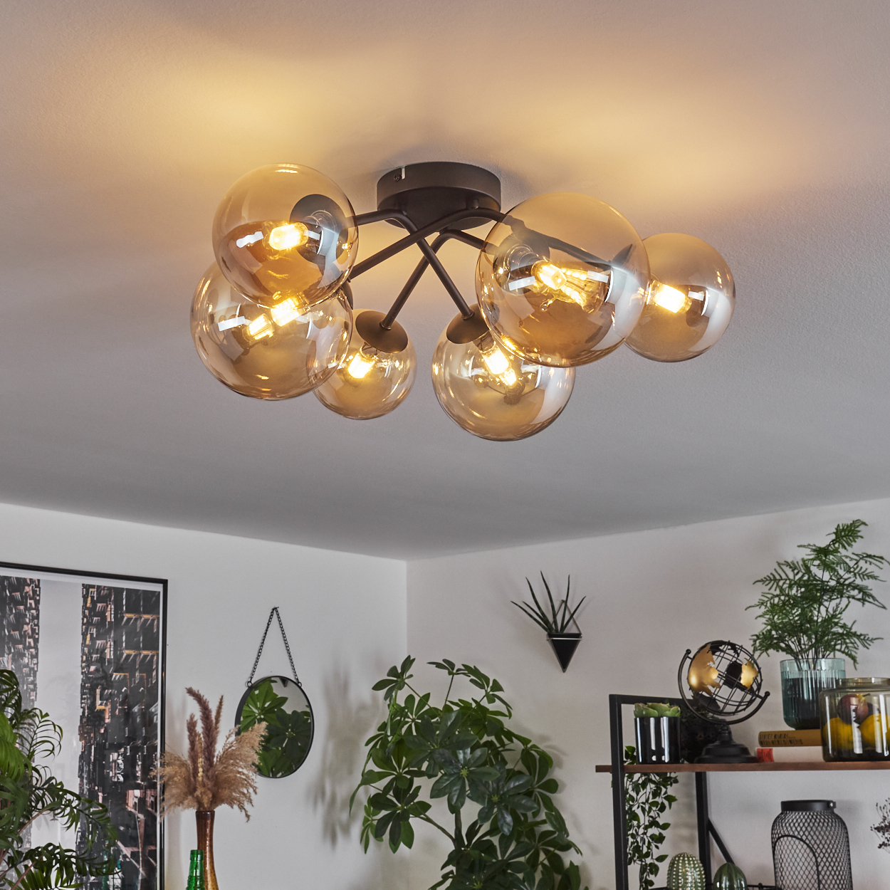 Deckenlampe mit bernsteinfarbenen Glaskugeln und schwarzem Rahmen, die einen Raum mit Heimdekor und Pflanzen beleuchtet.