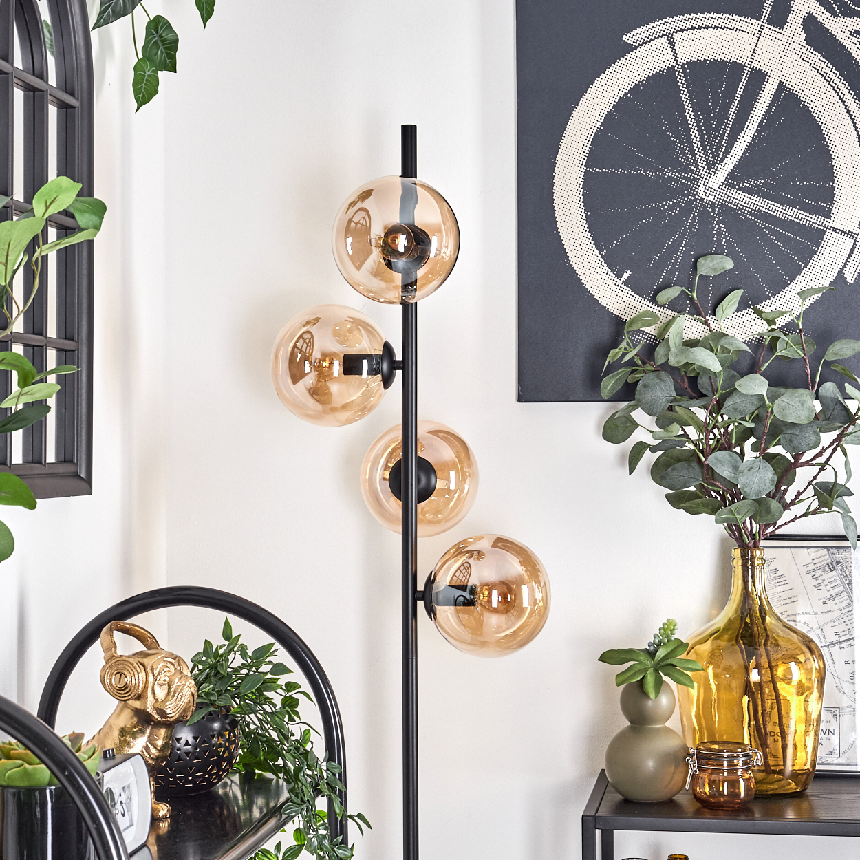 Schwarze Stehlampe mit vier bernsteinfarbenen Glaskugeln. Der Hintergrund umfasst Deko und einen Fahrrad-Kunstdruck.