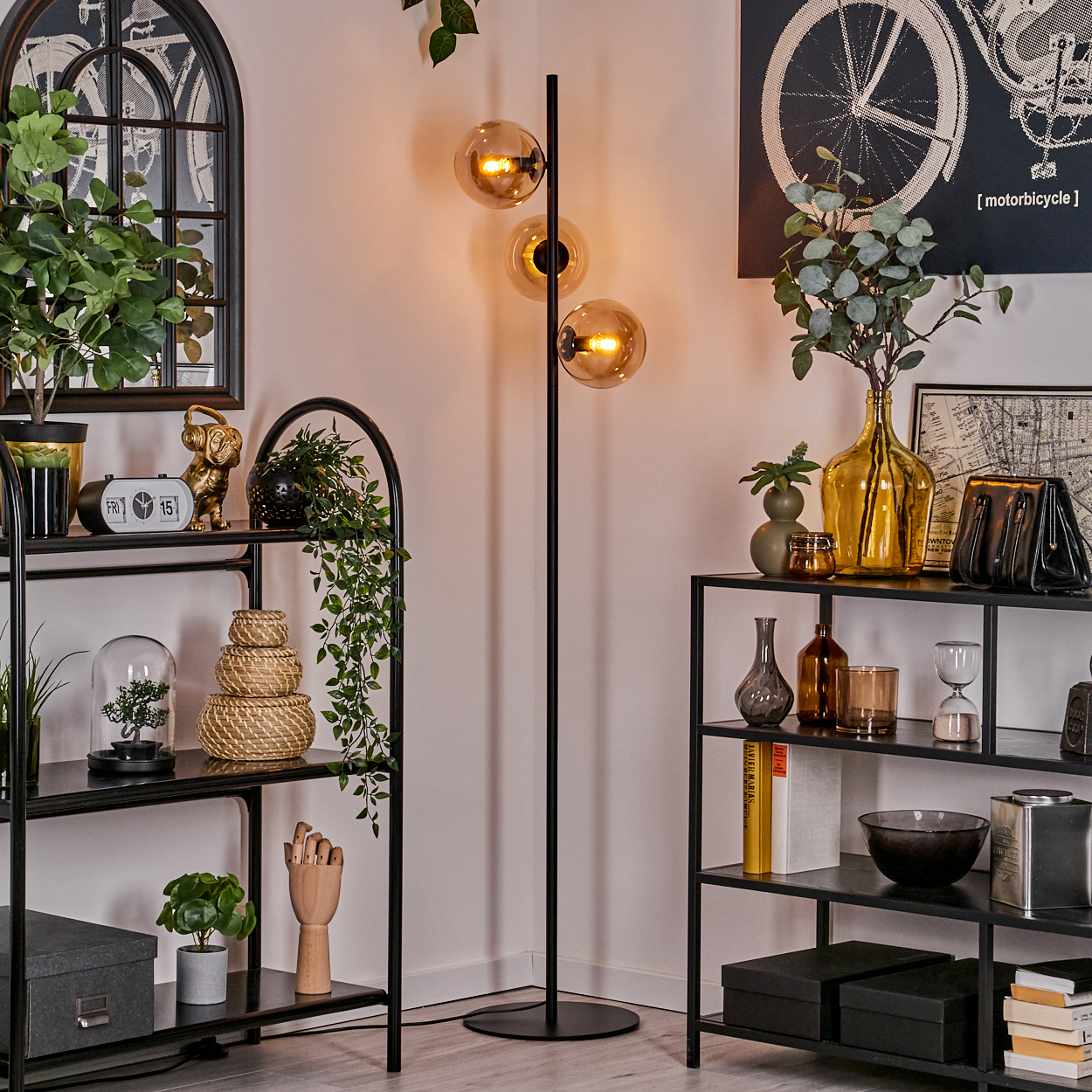 Stehlampe mit bernsteinfarbenen Glaskugeln, Dekor auf schwarzen Regalen und einem Fahrrad-Kunstdruck im Hintergrund.