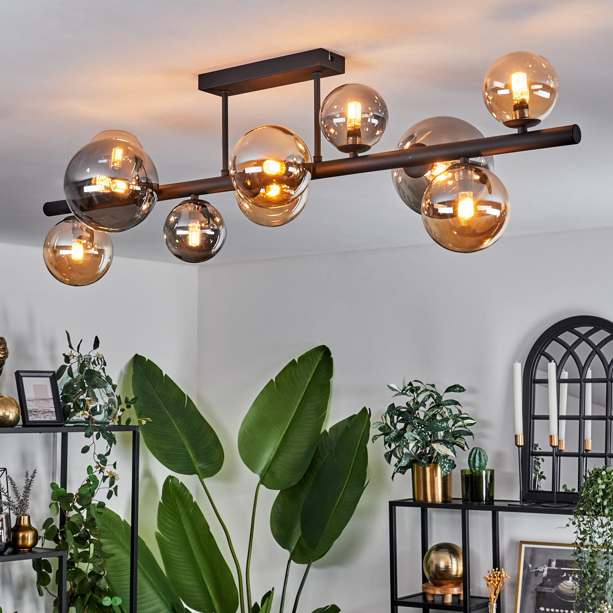 Deckenlampe mit acht bernsteinfarbenen und grauen Kugeln. Schwarzer Rahmen. Pflanzen im Hintergrund.