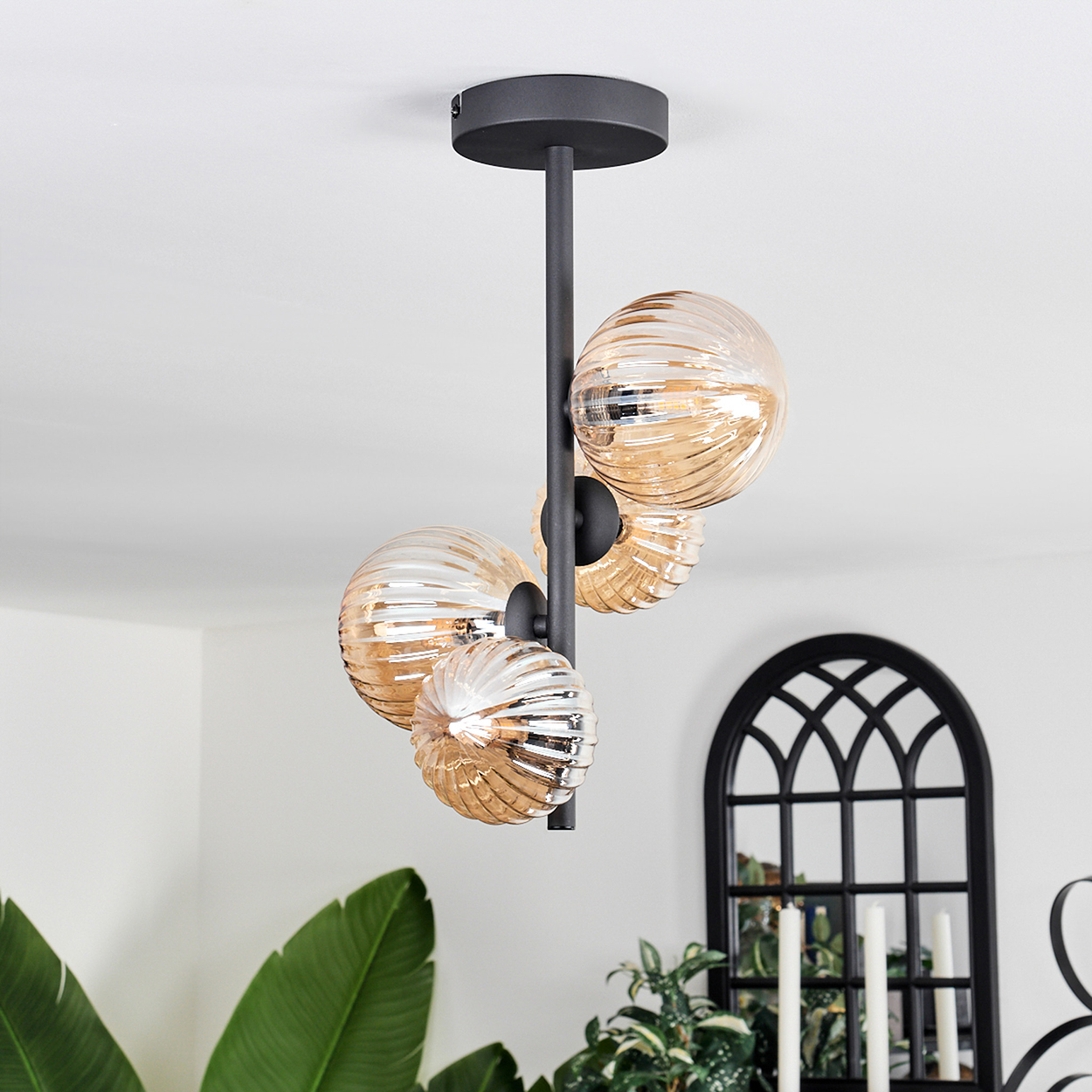 Deckenlampe mit bernsteinfarbenen Glaskugeln. Schwarzer Rahmen. Hintergrund mit Spiegel und Pflanzen. Weiße Decke.