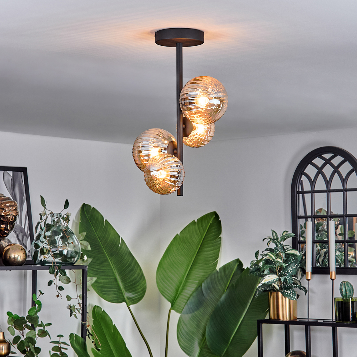 Deckenlampe mit bernsteinfarbenen Glaskugeln. Schwarzer Rahmen. Hintergrund mit Pflanzen, Spiegel und schwarzem Regal.