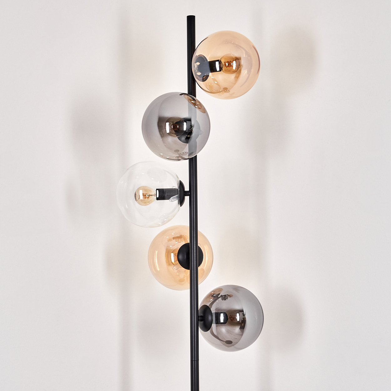 Eine schwarze Stehlampe mit fünf Kugeln aus bernsteinfarbenem und silbernem Glas vor einer weißen Wand.