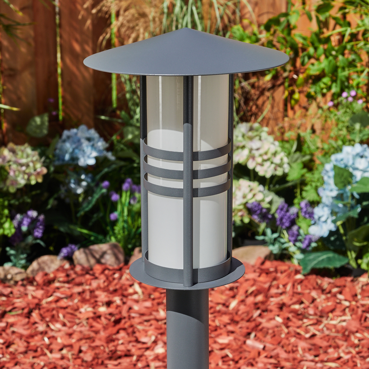 Eine Außenlaterne mit zylindrischem weißem Licht und flachem Dach, in einem Garten.