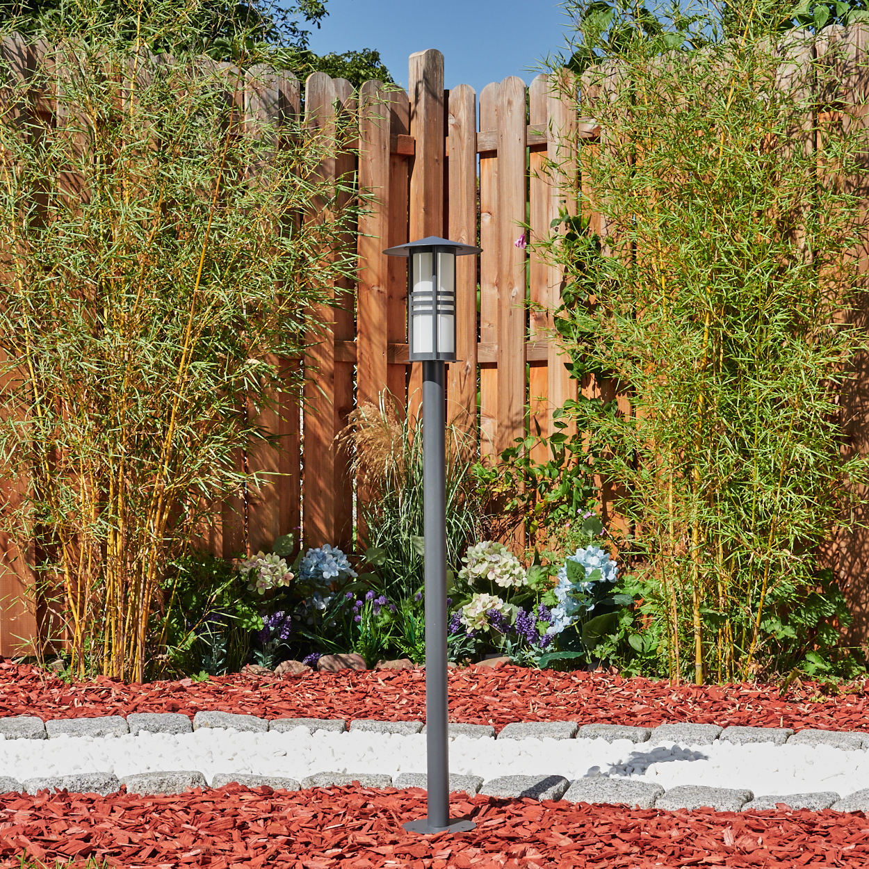 Außen-Laternenpfahl im Garten. Grauer Metallpfahl, weißes Licht. Bambus und Holzzaun im Hintergrund.
