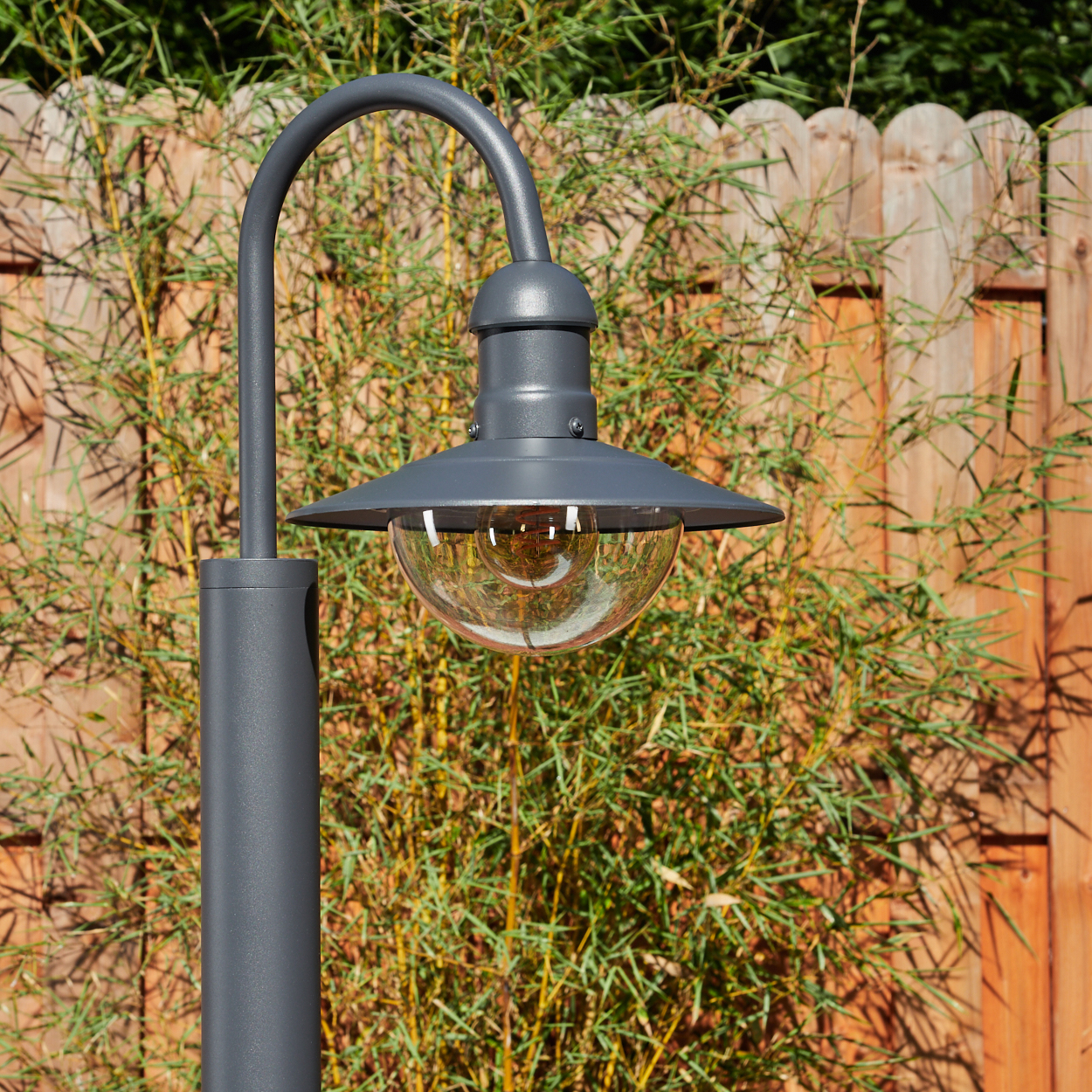 Eine graue Außenlampe mit gebogenem Arm und Halbkugel-Schirm, vor einem Hintergrund aus Holzzaun.