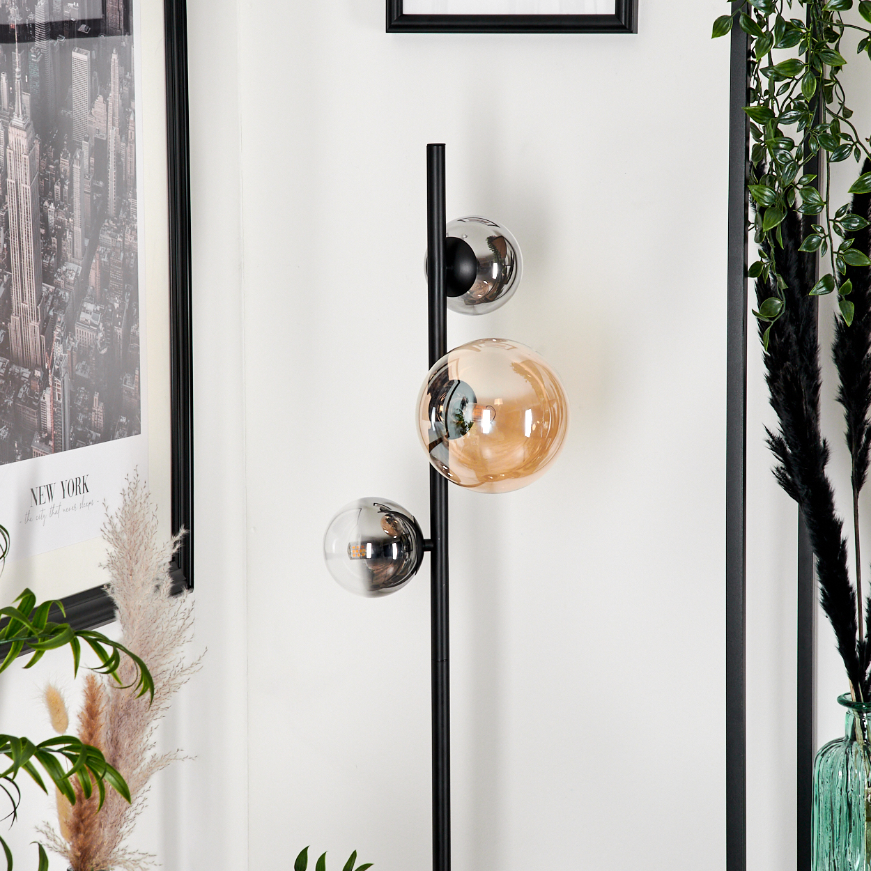 Schwarze Stehlampe mit drei bernsteinfarbenen Glasschirmen. Die Lampe steht neben einer Wand.