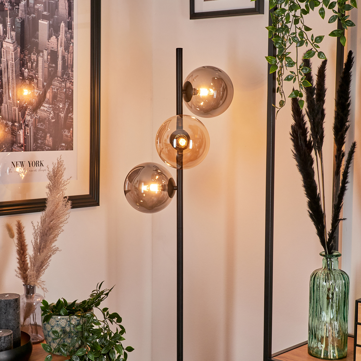 Stehlampe mit drei Kugelleuchten in Bernstein neben New York-Druck, Vase und Pflanzen.