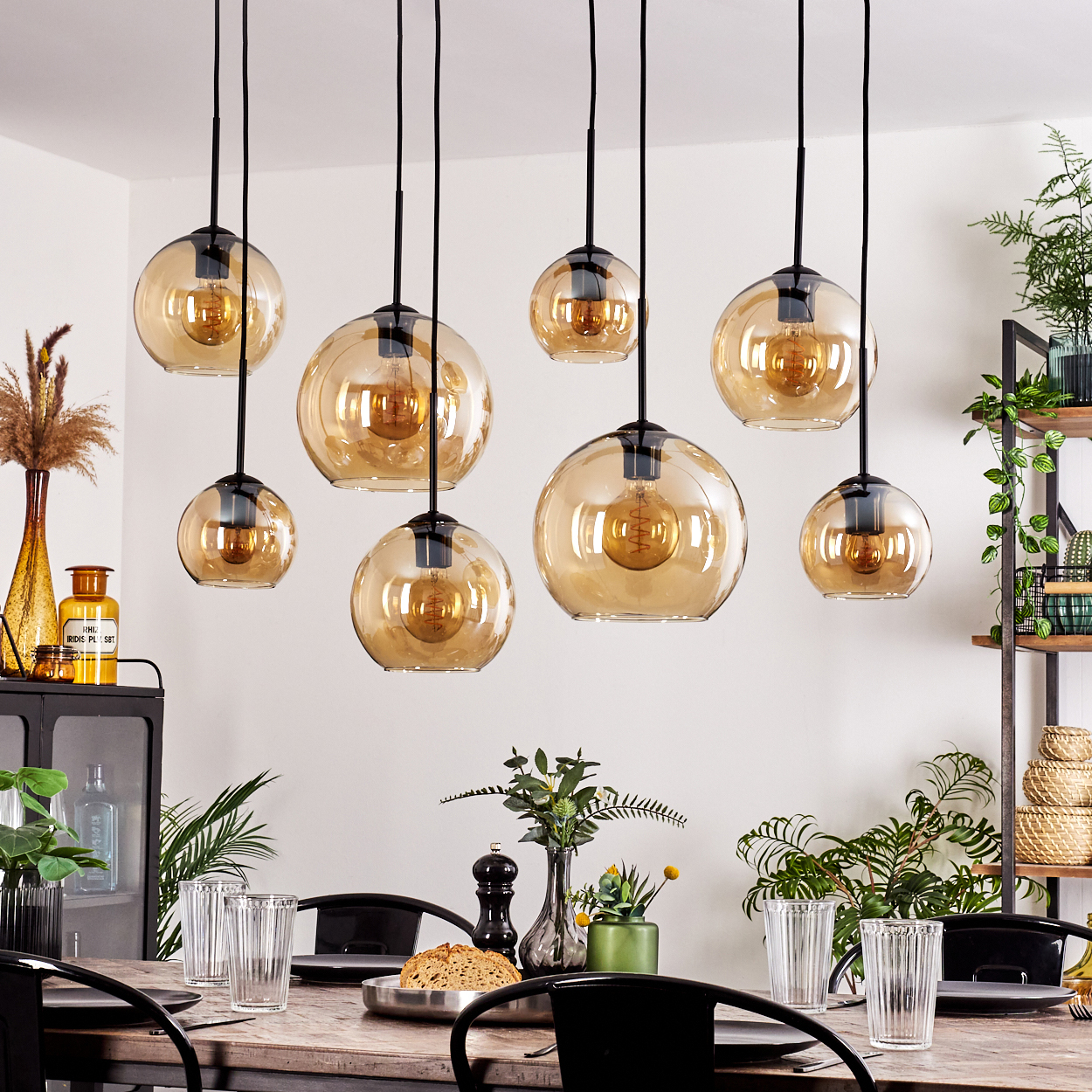 Acht Pendelleuchten aus bernsteinfarbenem Glas hängen über einem Tisch. Pflanzen, Gläser und Dekoration im Hintergrund.