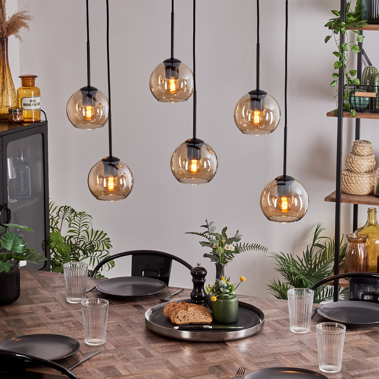 Sechs Pendelleuchten mit bernsteinfarbenen Glaskugeln über einem Esstisch. Teller, Gläser und Essen sind auf dem Tisch.