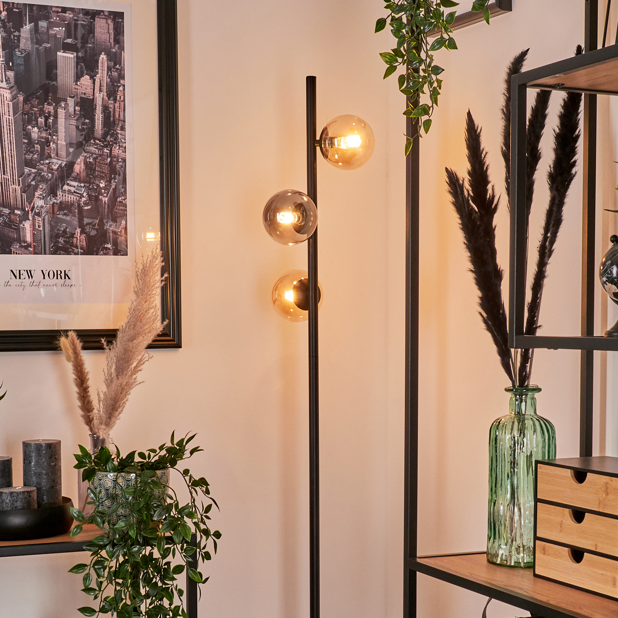 Stehlampe mit drei bernsteinfarbenen Glaskugeln. Pflanzen und Regale sind im Hintergrund. Ein New York-Poster an der Wand.