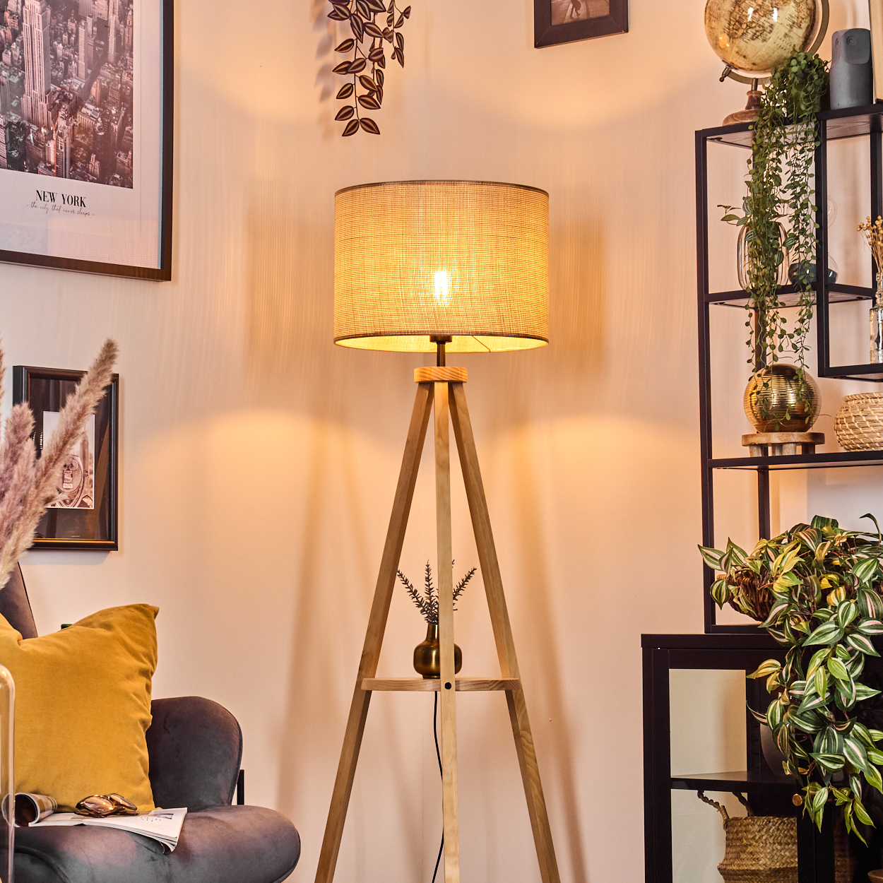 Eine beleuchtete Holz-Stativlampe steht in einem Raum. Die Lampe hat einen beigefarbenen Schirm und beleuchtet den Raum.