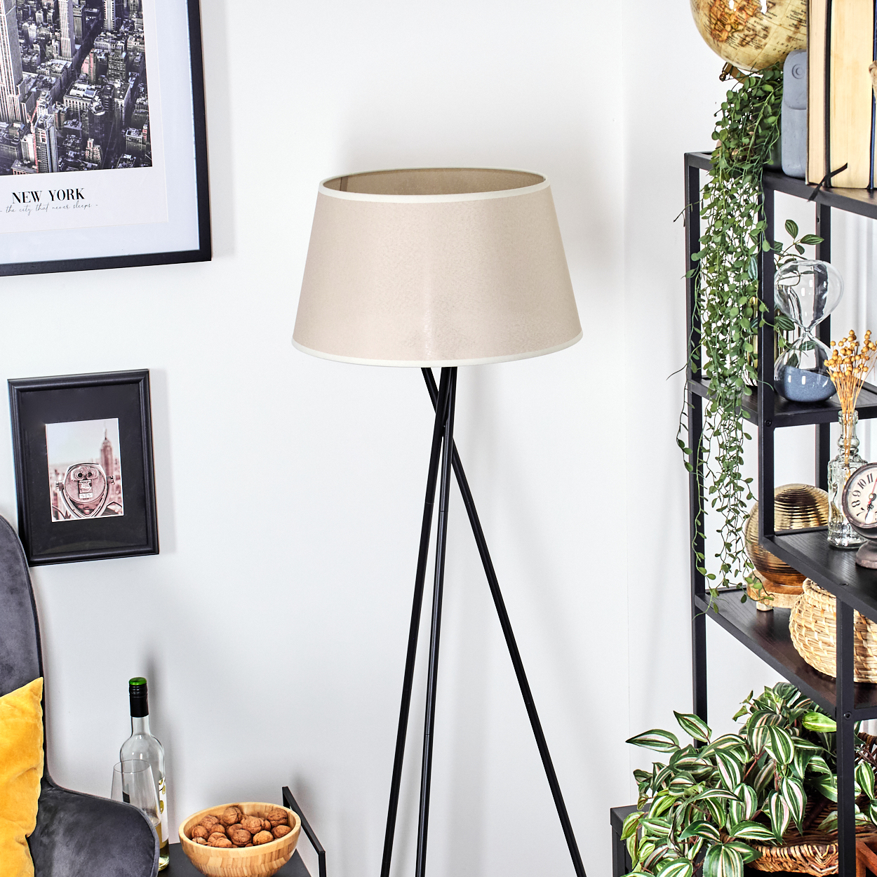 Dreibein-Stehlampe mit beigem Schirm, schwarzen Beinen und hellem Licht. Neben Regal.