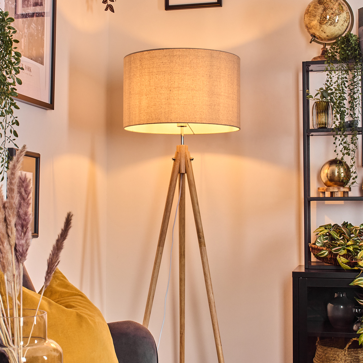 Dreibein-Stehlampe in einem Raum. Die Lampe hat einen beigen Lampenschirm, helle Holzbeine und ist beleuchtet.