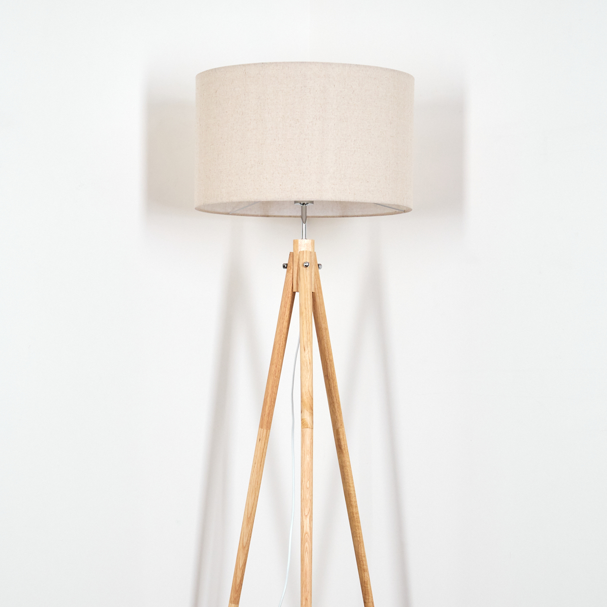 Eine Holz-Stativ-Stehlampe mit beigem Lampenschirm steht vor einer weißen Wand.