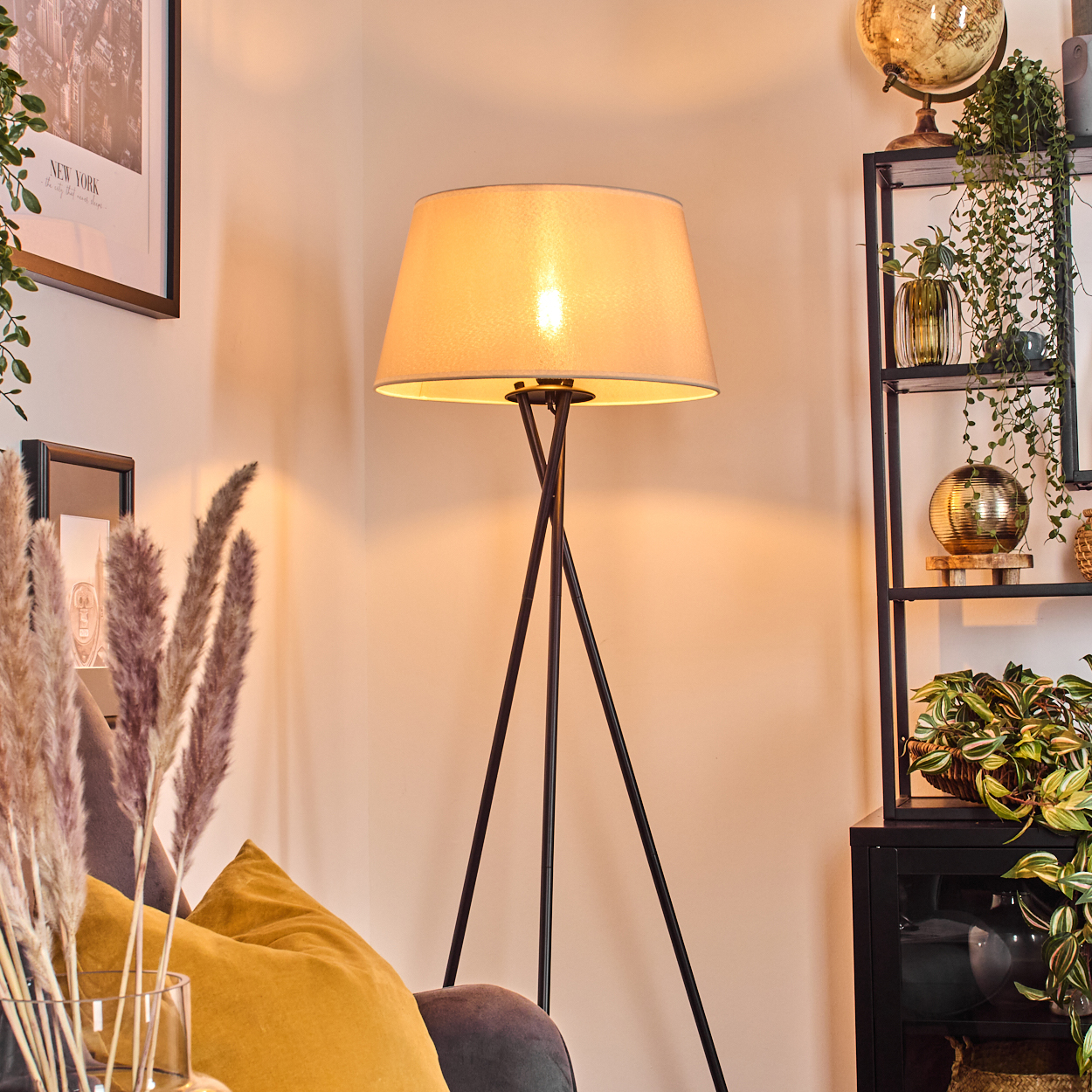 Dreibein-Stehlampe in einem Raum. Die Lampe hat einen beigen Lampenschirm, schwarze Beine und ist beleuchtet.