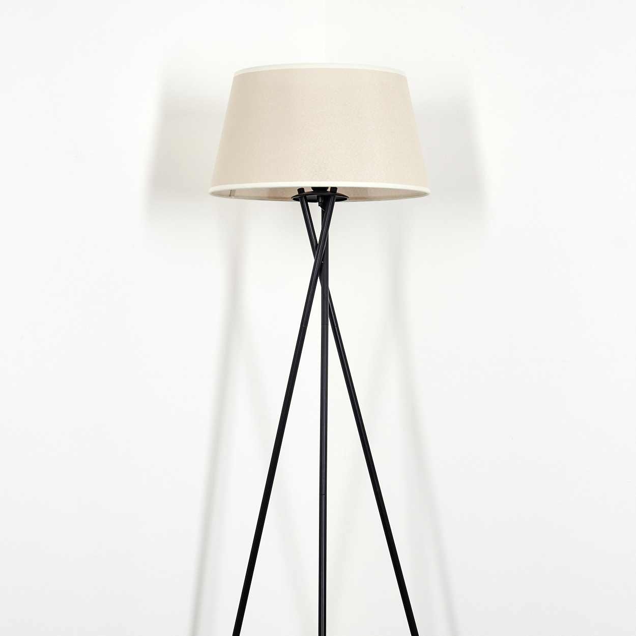Eine Stehlampe mit beigem Lampenschirm und schwarzen Beinen, vor einer weißen Wand.
