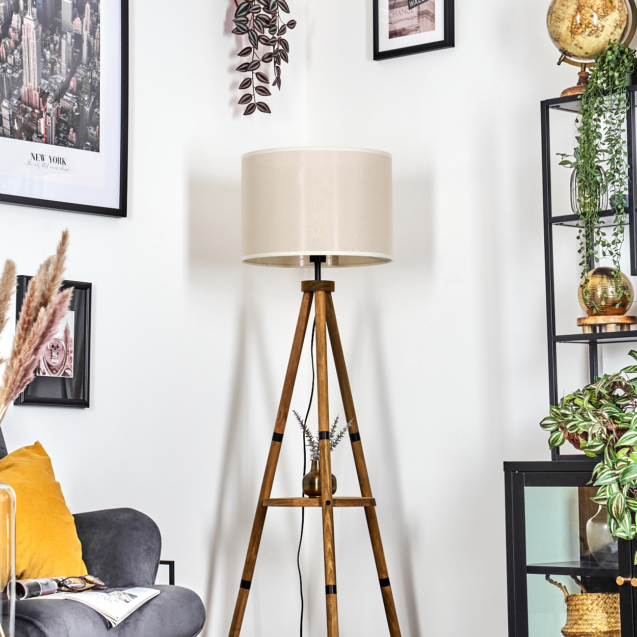 Eine Holzstativlampe mit weißem Lampenschirm steht neben einem grauen Sessel und gerahmten Bildern.