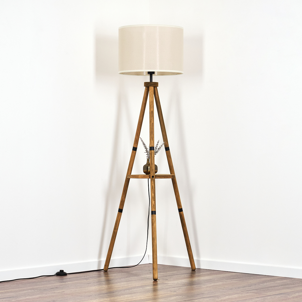 Eine Holzstativlampe mit beigem Lampenschirm steht in einer Ecke vor weißer Wand und Holzboden.