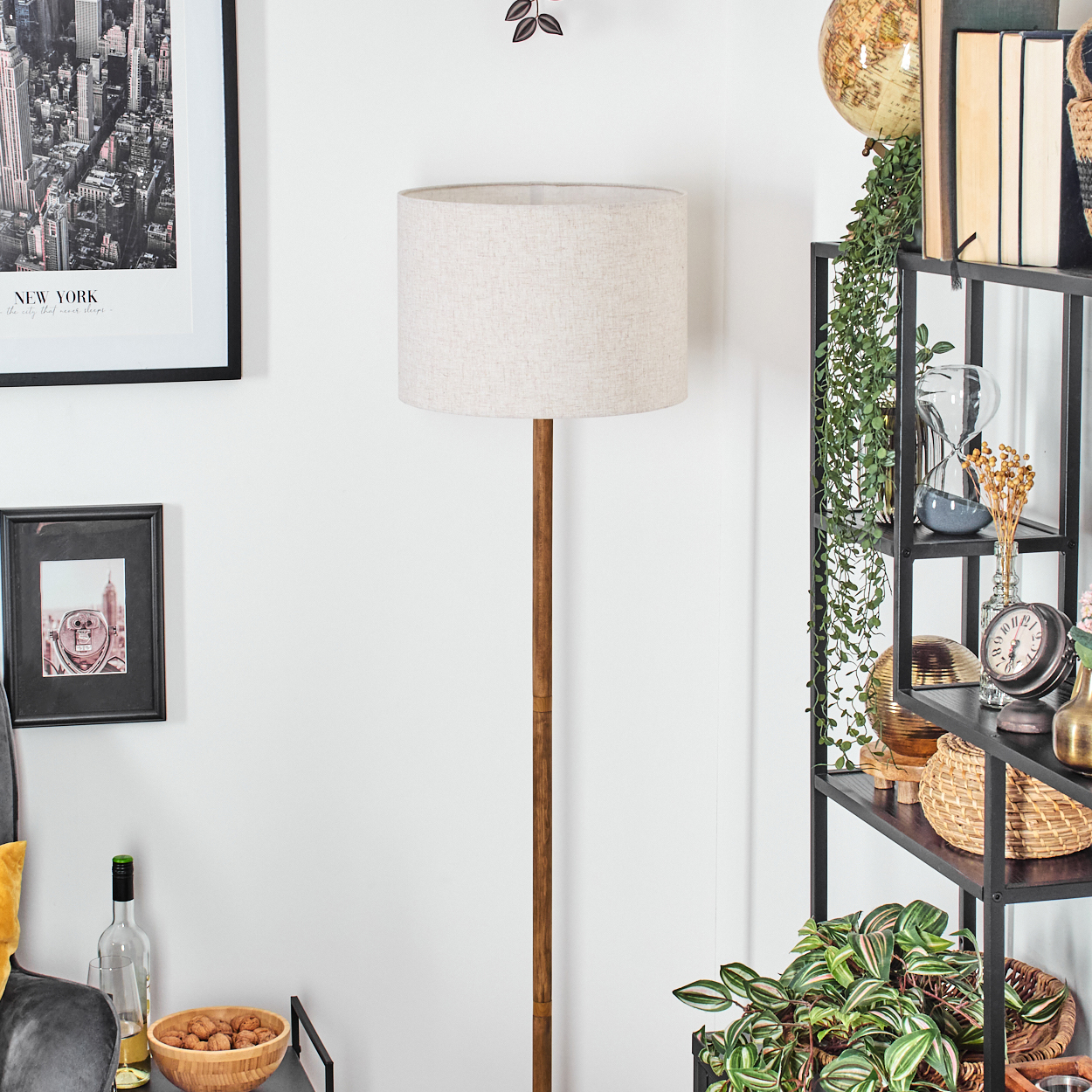 Eine Stehlampe mit beigem Schirm, Holzstange und einem Regal mit Gegenständen und Pflanzen.
