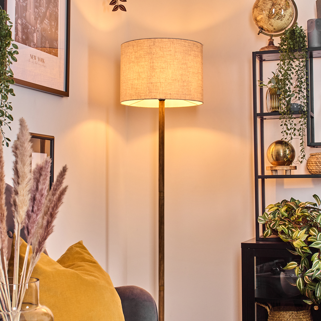Eine hohe Stehlampe mit beigem Lampenschirm. Ein gelbes Kissen und eine Pflanze sind sichtbar.