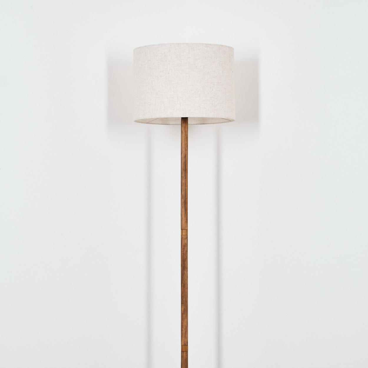 Hohe Stehlampe aus Holz. Beiger Lampenschirm oben. Braune Stange und weißer Wandhintergrund.
