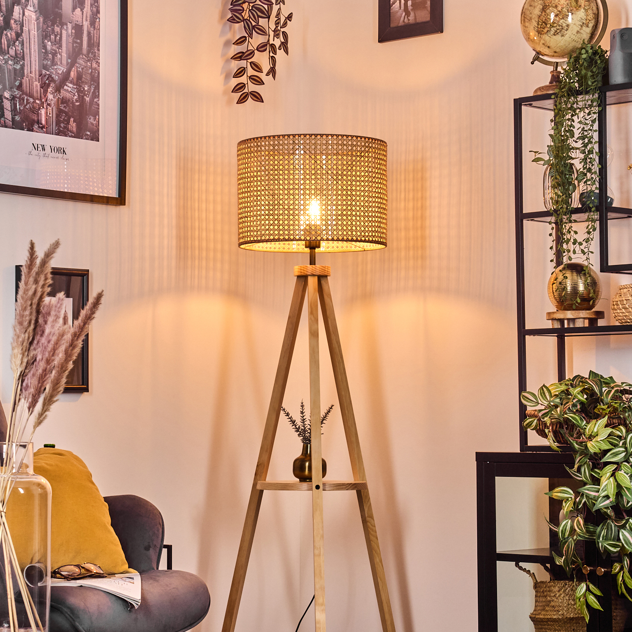 Dreibeinlampe in einem Raum. Rattan-Schirm, New York Poster, Regal mit Deko. Warmes Licht.
