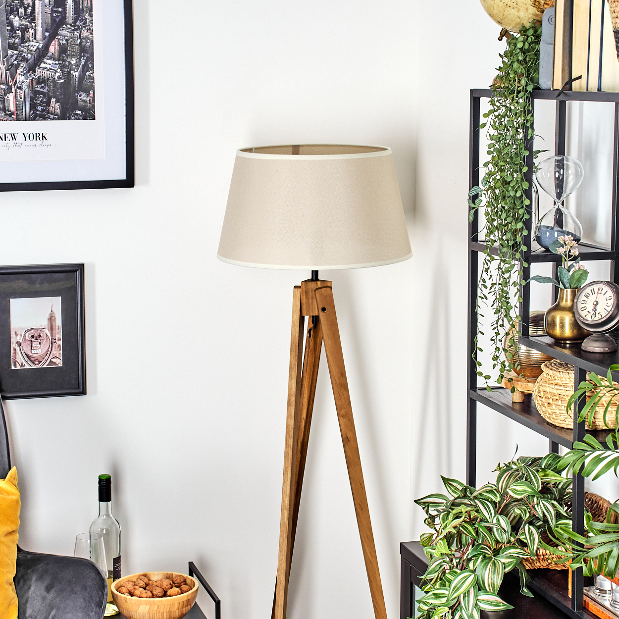 Stativ-Stehleuchte aus Holz mit beigem Lampenschirm, neben einer Wand mit Kunst und Regalen.