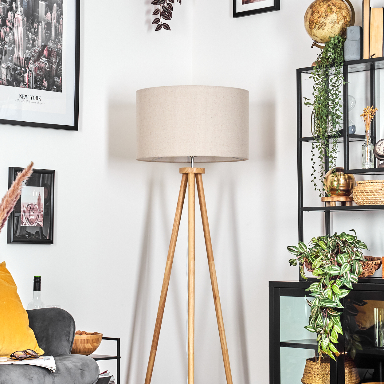 Hölzerne Stativlampe mit beigem Lampenschirm. Sofa, Bilder und Pflanzen im Hintergrund.