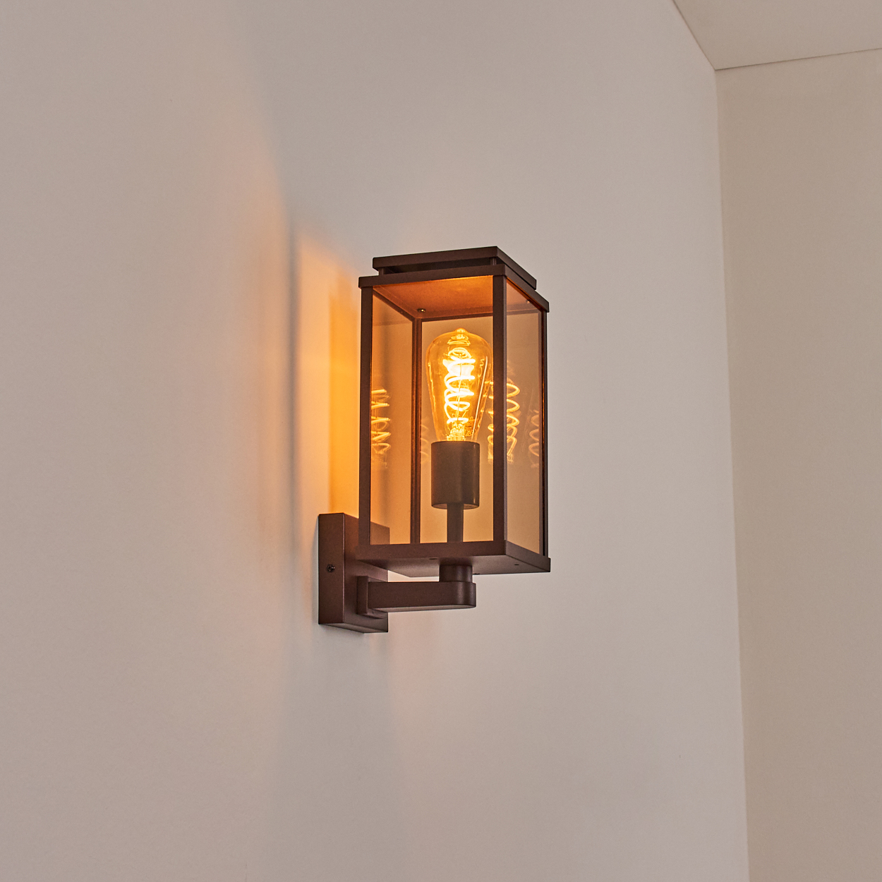 Bronze-Außenwandleuchte mit Glasscheiben und einer leuchtenden Glühbirne, an weißer Wand.