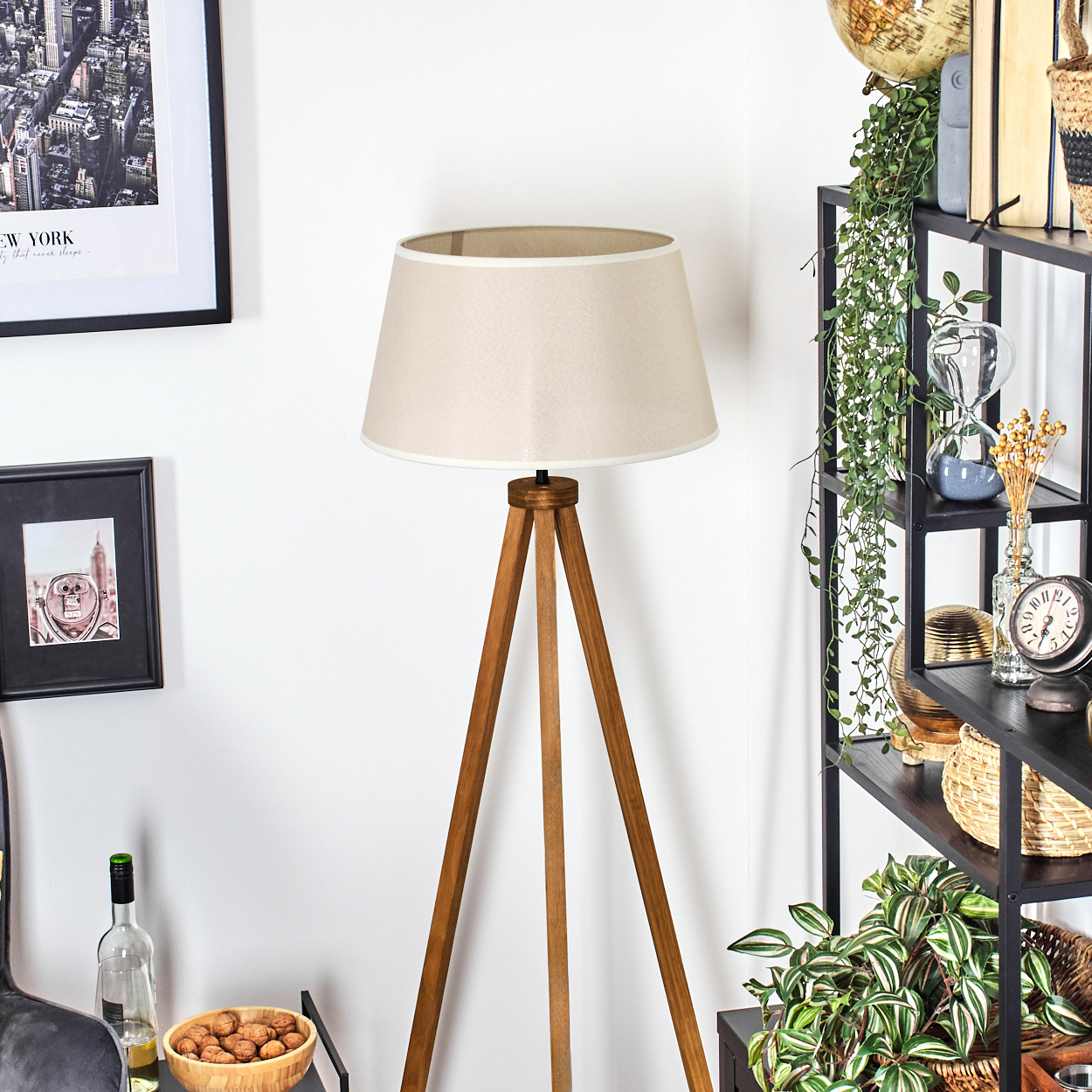 Stehlampe mit beigem Schirm und Holz-Dreibein. Raum mit Deko: Bilder, Regale.