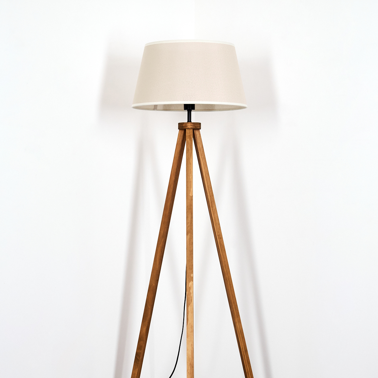 Eine hölzerne Stativ-Stehlampe mit beigem Schirm vor weißer Wand.