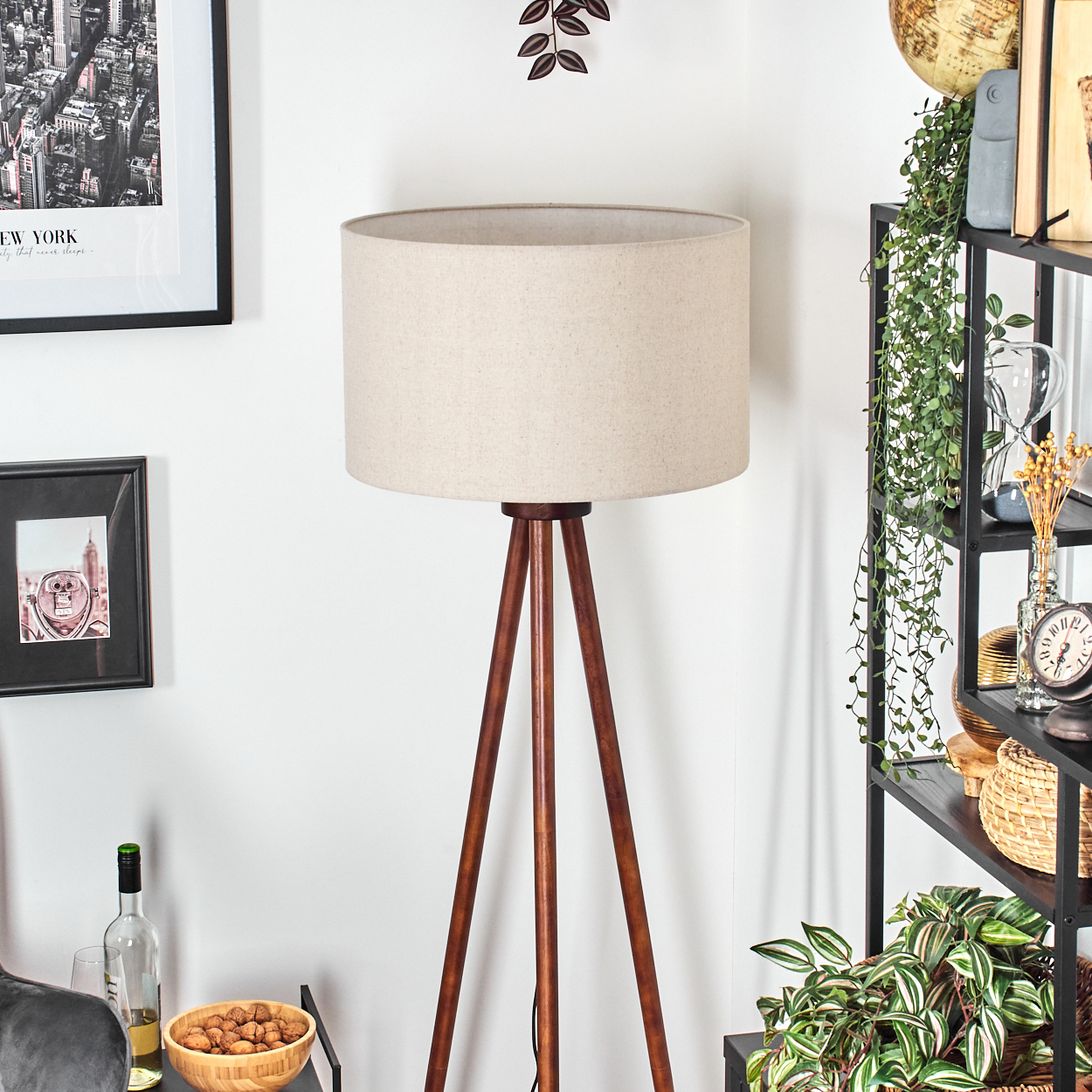 Eine hölzerne Stativlampe mit beigem Schirm in der Nähe einer Wand mit Deko.
