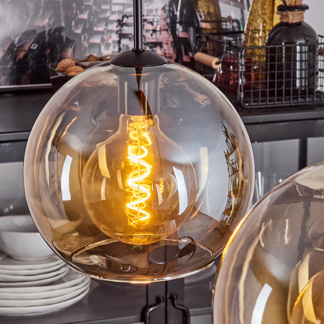 Eine Glas-Pendelleuchte mit sichtbarer Glühbirne. Hintergrund: Regale und Gegenstände.