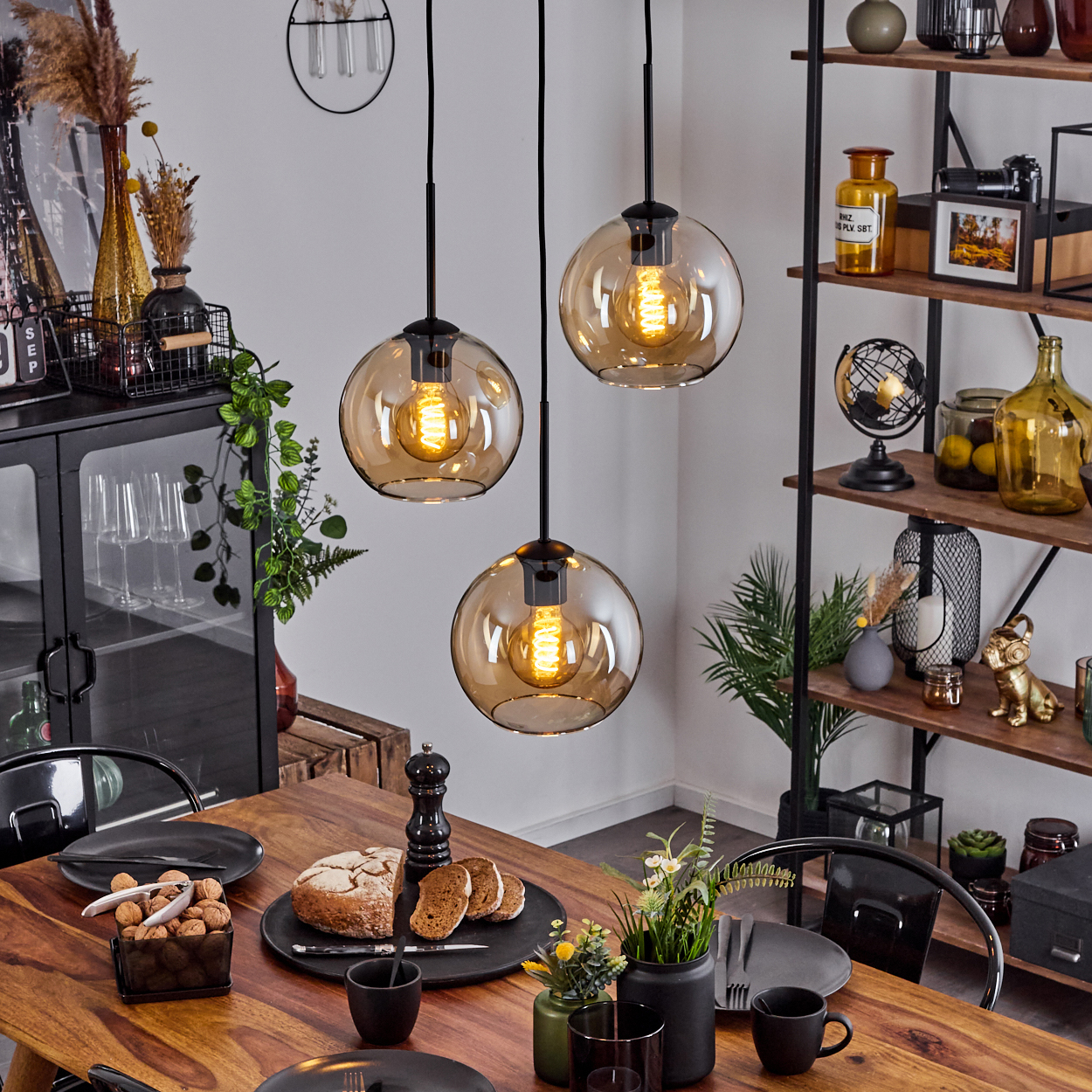 Drei Pendelleuchten über einem Holztisch. Bernsteinfarbene Glasschirme, schwarze Armaturen. Regale.