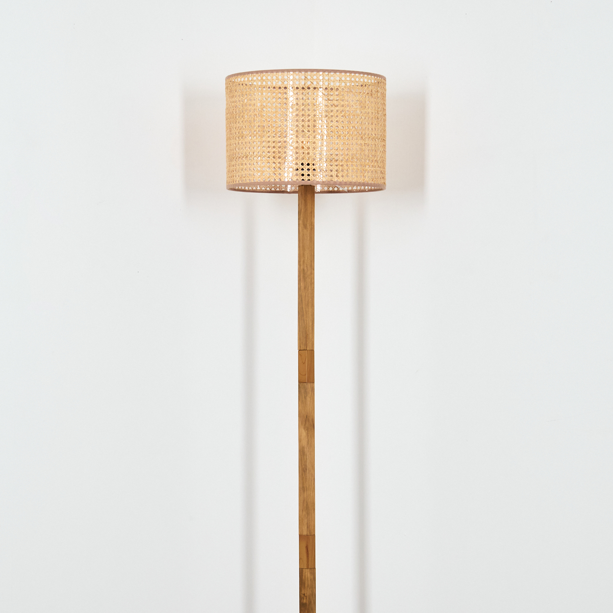 Eine hohe Stehlampe mit gewebtem Lampenschirm vor einer weißen Wand. Die Lampe ist aus Holz gefertigt.