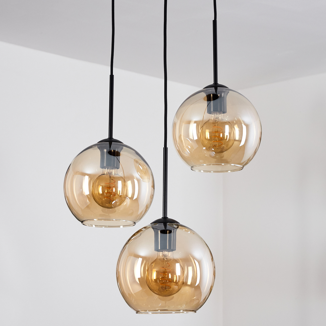 Drei Pendelleuchten mit freiliegenden Glühbirnen vor einer weißen Wand. Lampen haben schwarze Fassungen und Glaskugeln.
