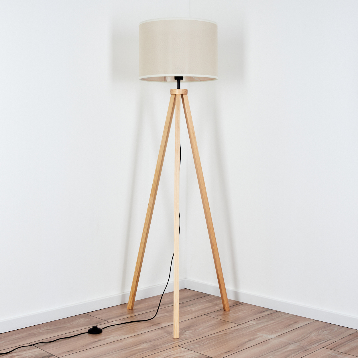 Stativlampe in einer Ecke. Beiger Lampenschirm, Holzbeine, schwarzes Kabel und Schalter auf Holzboden.