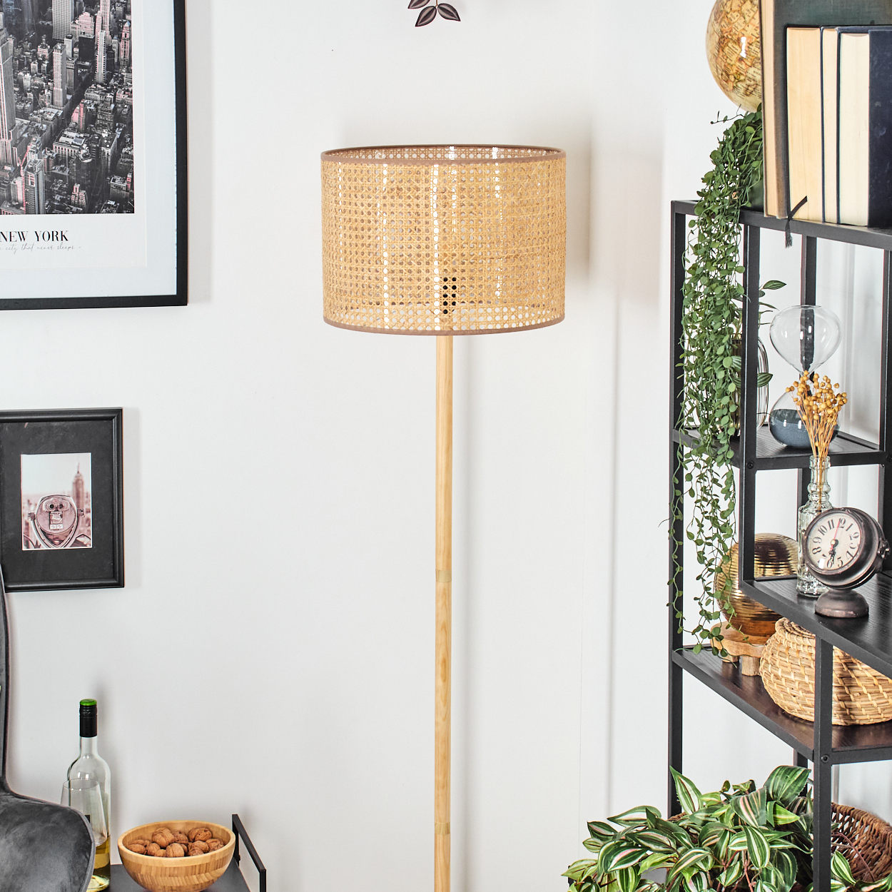Stehlampe mit gewebtem beigefarbenem Schirm, in einem Raum mit einem Gemälde, einem Regal und Pflanzen im Hintergrund stehend.