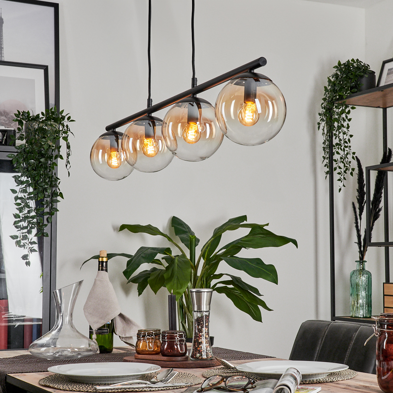 Schwarze Deckenlampe mit vier bernsteinfarbenen Glaskugeln. Die Szene beinhaltet einen Esstisch mit Gläsern und Pflanzen.