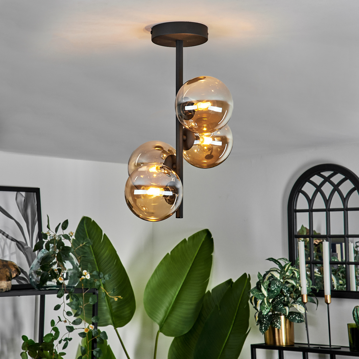 Schwarze Deckenlampe mit vier bernsteinfarbenen Glaskugeln. Die Szene umfasst Pflanzen und Dekorationen im Hintergrund.