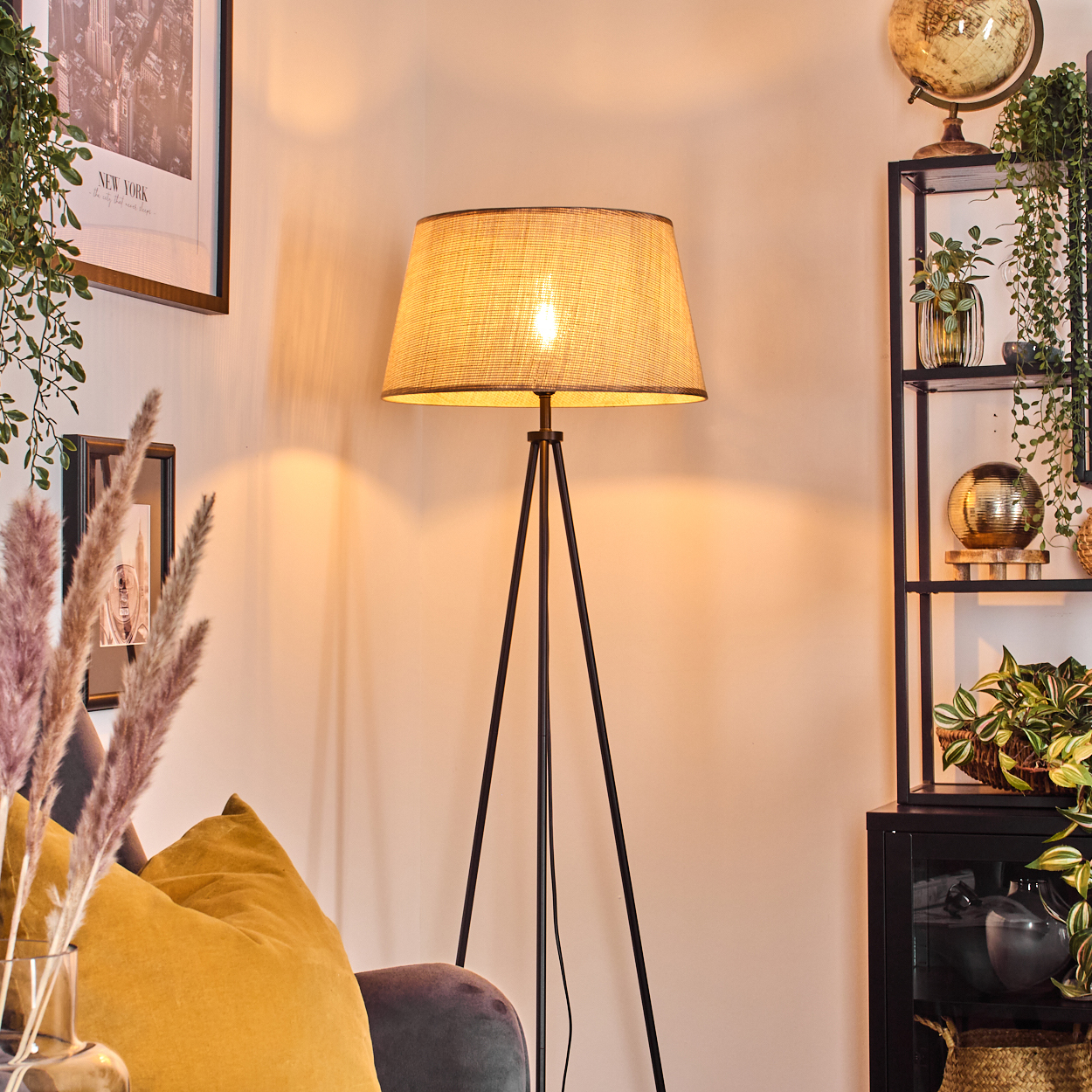 Dreibein-Stehlampe mit Lampenschirm, beleuchtete Glühbirne. Innenraum mit Regalen und Pflanzen.
