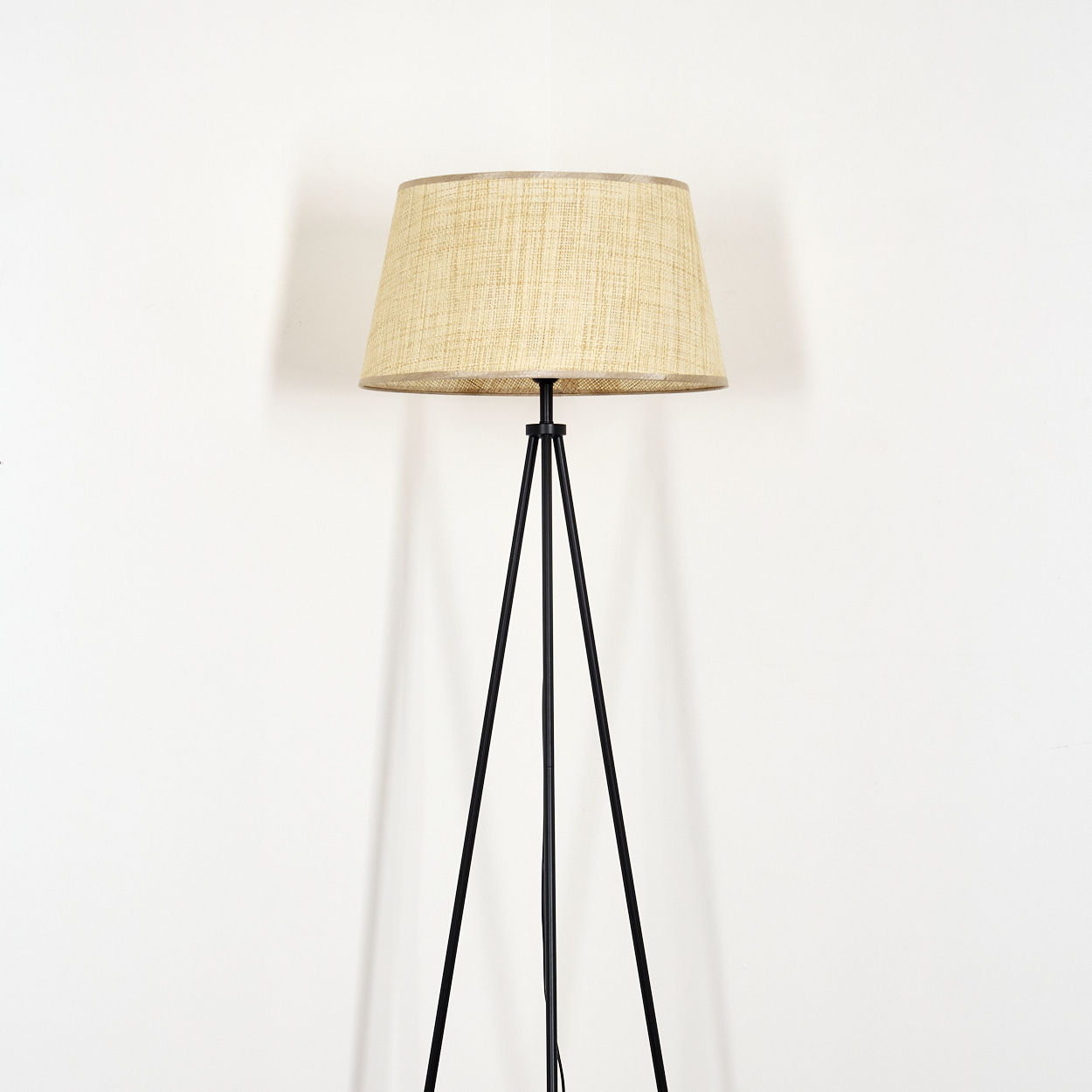 Eine einfache Stehlampe mit beigem Lampenschirm und schwarzen Metallbeinen, vor weißem Hintergrund.