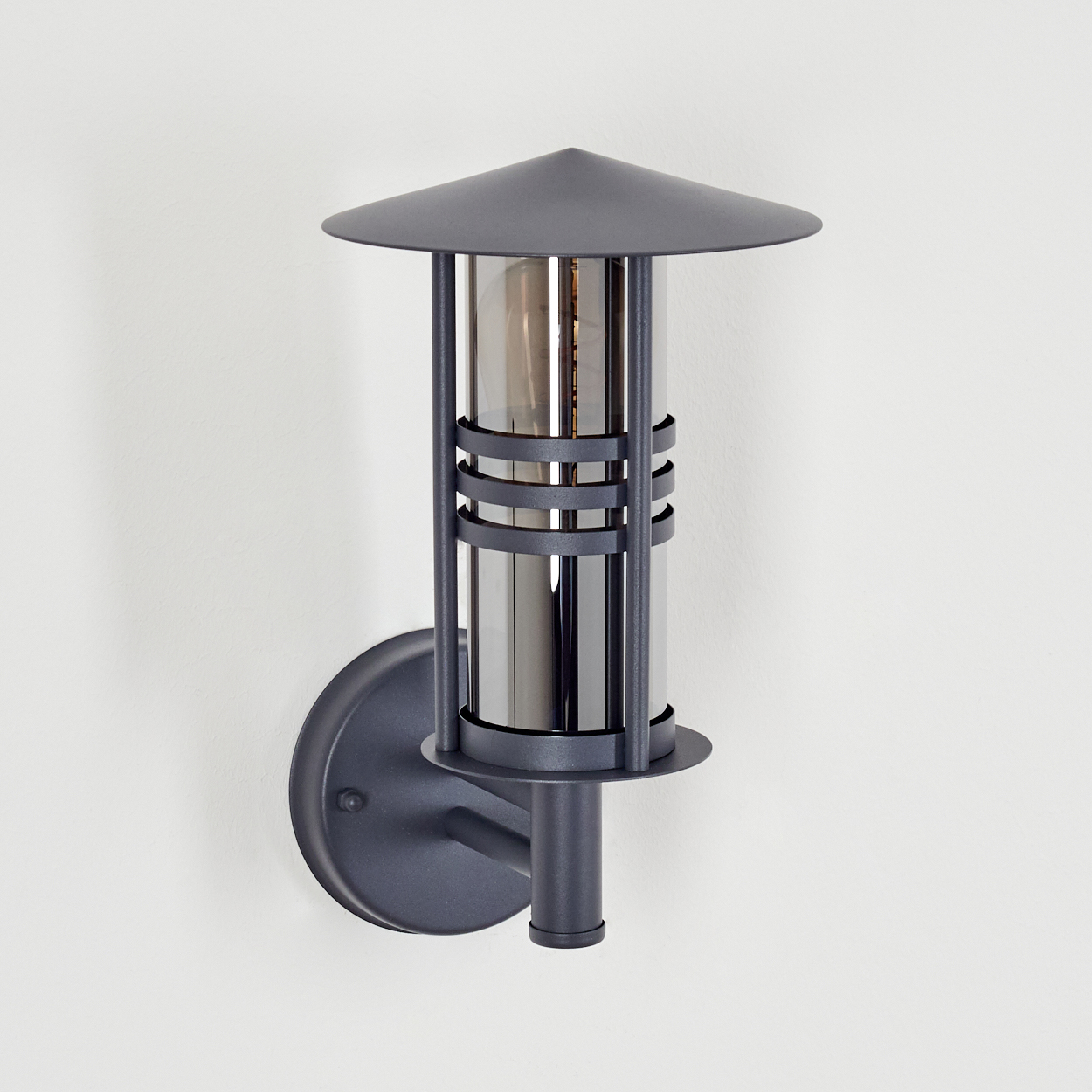 Eine graue Außenlampe an einer weißen Wand, mit zylindrischem Design und kleinem Schirm.